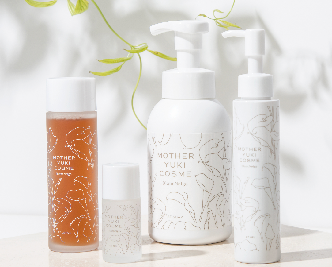 Four natural skincare products from the Mother Yuki Cosme brand, displayed on a white surface with green leaves in the background. The products include a cleansing oil, a face lotion, and two other skincare items, all with minimalist white and printed designs.