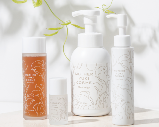 Four natural skincare products from the Mother Yuki Cosme brand, displayed on a white surface with green leaves in the background. The products include a cleansing oil, a face lotion, and two other skincare items, all with minimalist white and printed designs.